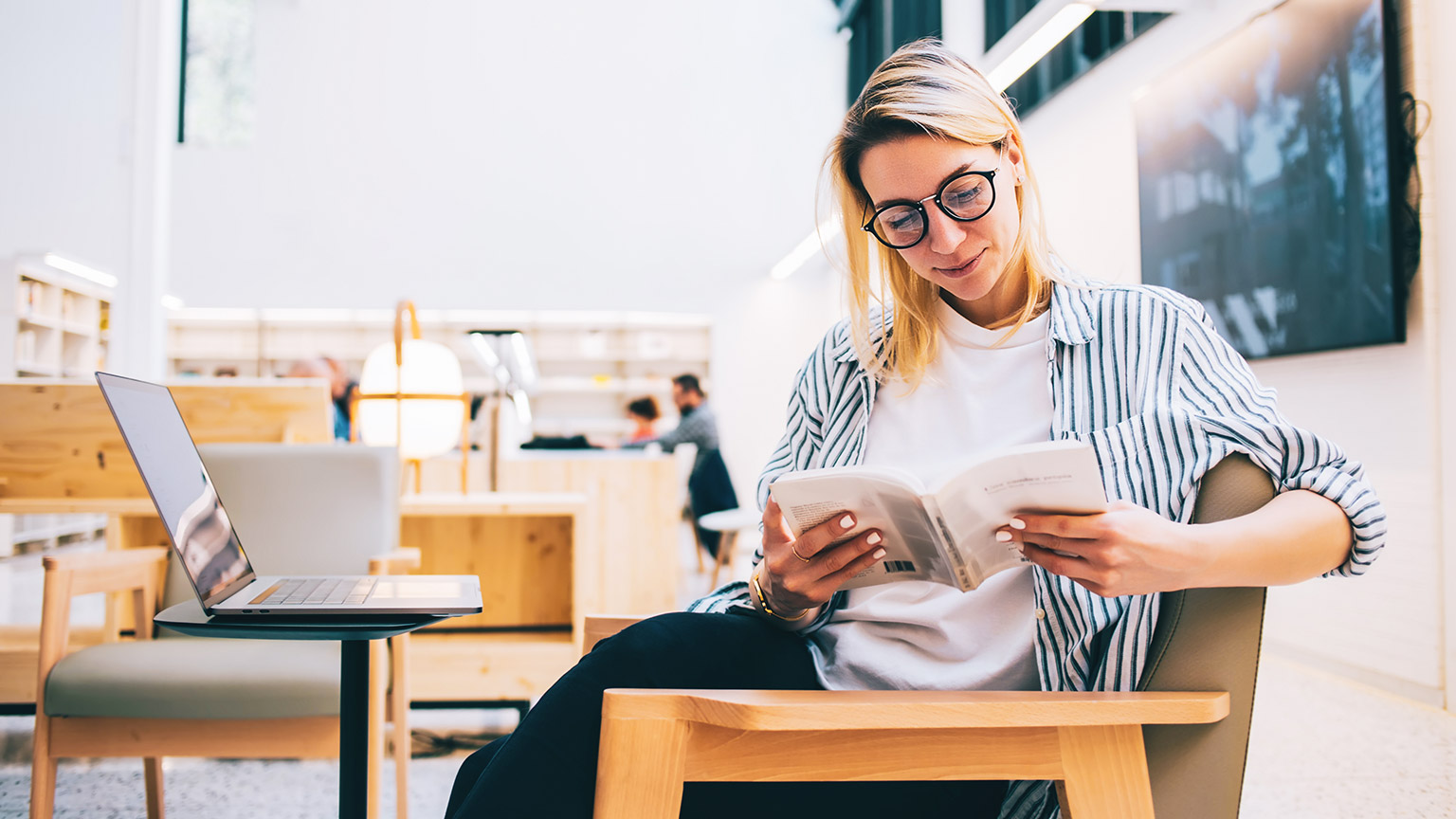 Women in library reading a book with laptop on the table beside her