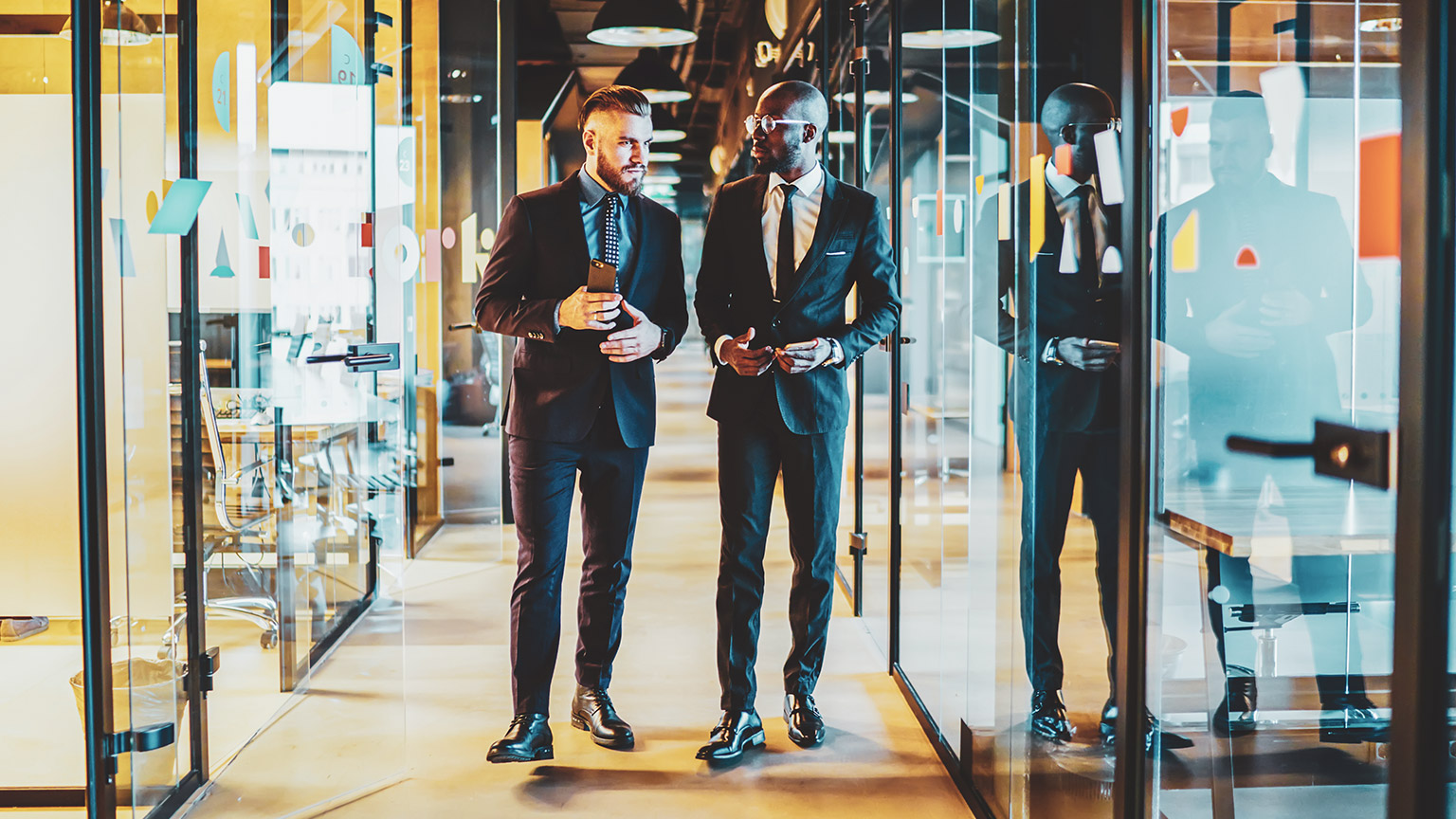 2 males in business attire walking down hallway of modern office while talking