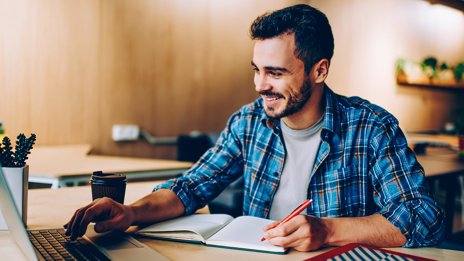 Young male hipster with a wry smile sitting in a library using a laptop while writing in a notepad