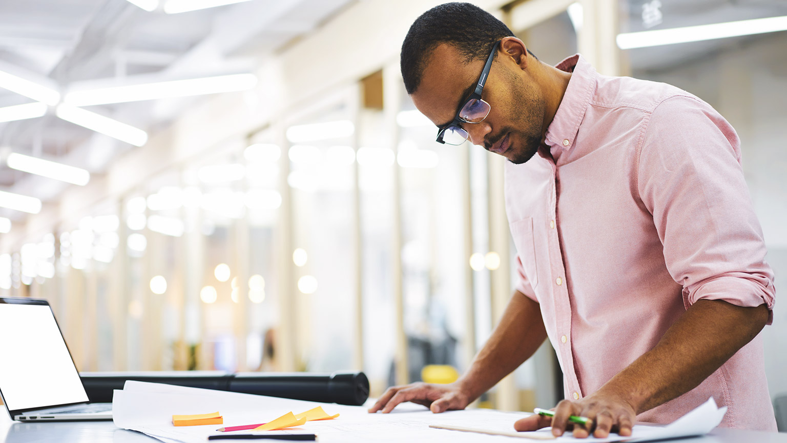 Male construction manager with glasses standing at a desk in a bright office looking at plans on desk and about to make amendments to them