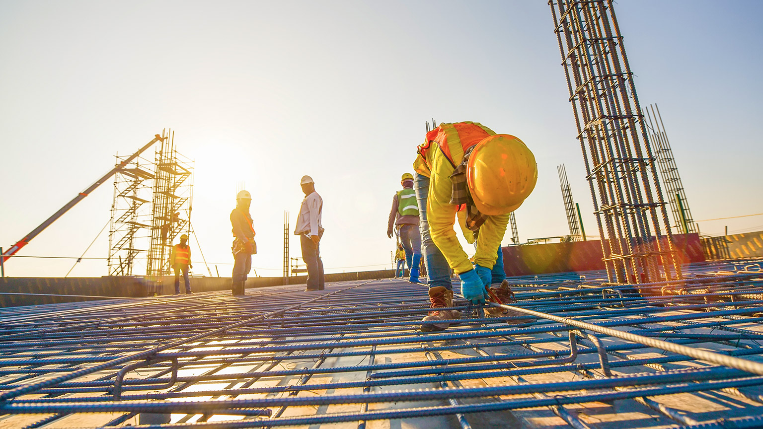 A wide shot of a construction worker tying down reinforced steel, with other workers and machinery in the background