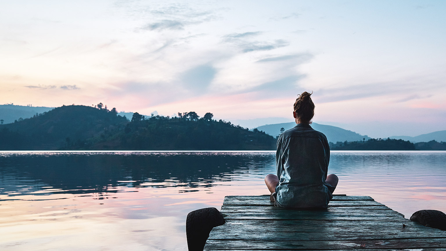 A young woman sitting alone on a pier looking over a calm lake at sunset