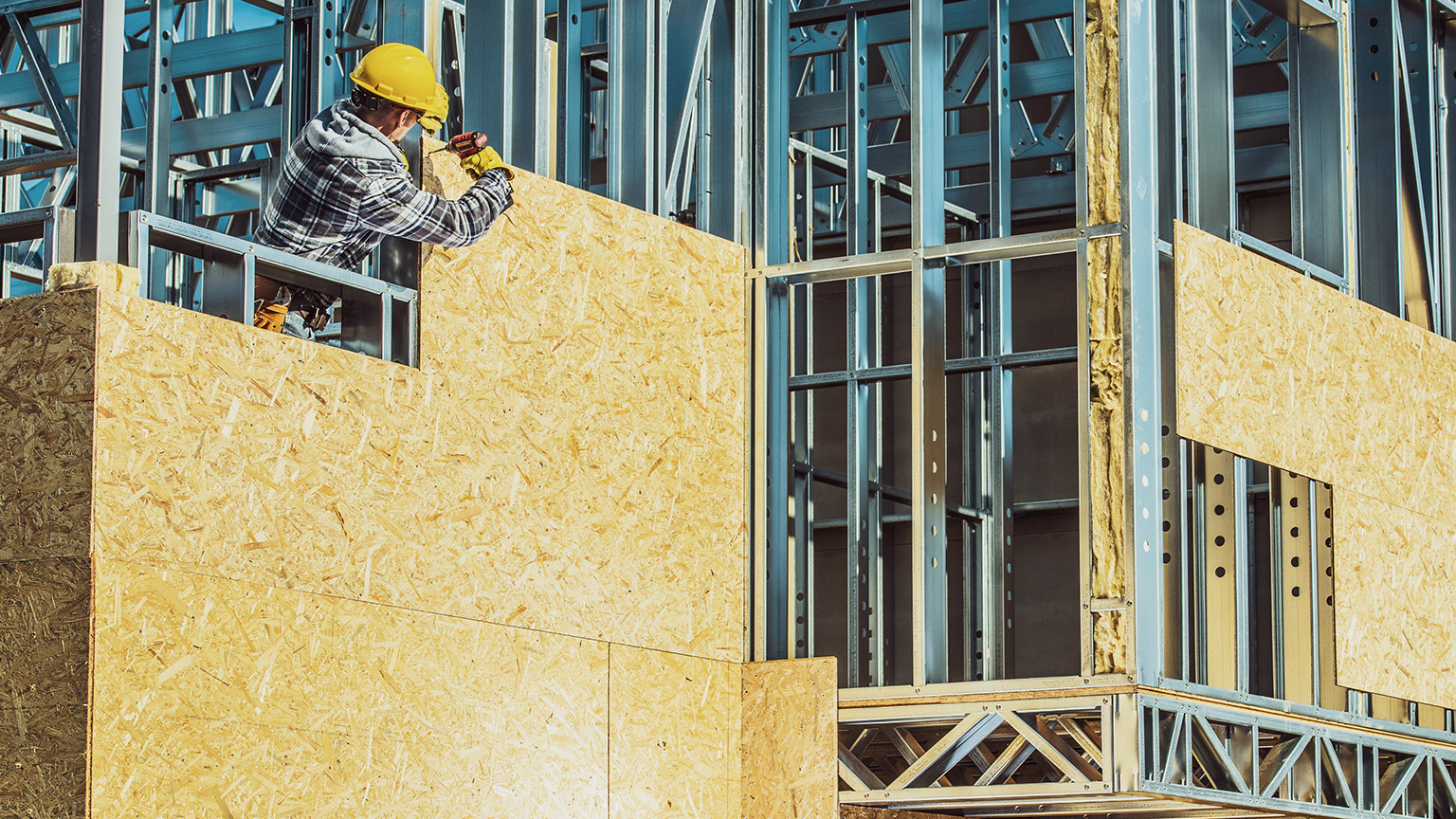 A wide view of a construction site with a worker attaching plywood to a frame