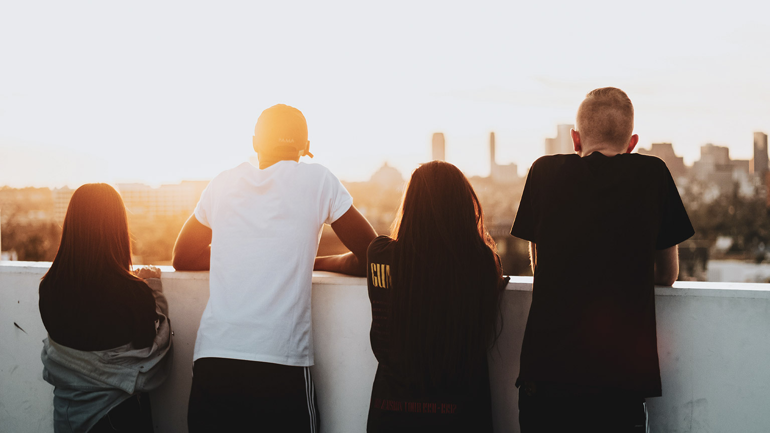 A diverse group of young people with back to camera looking out over a city at sunset