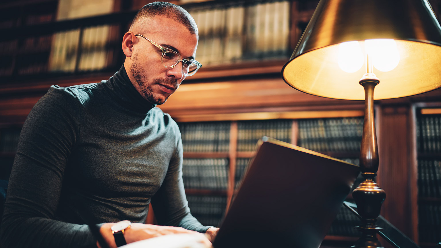 An academically-minded hipster sitting in a rich, mahogany library working on a writing project