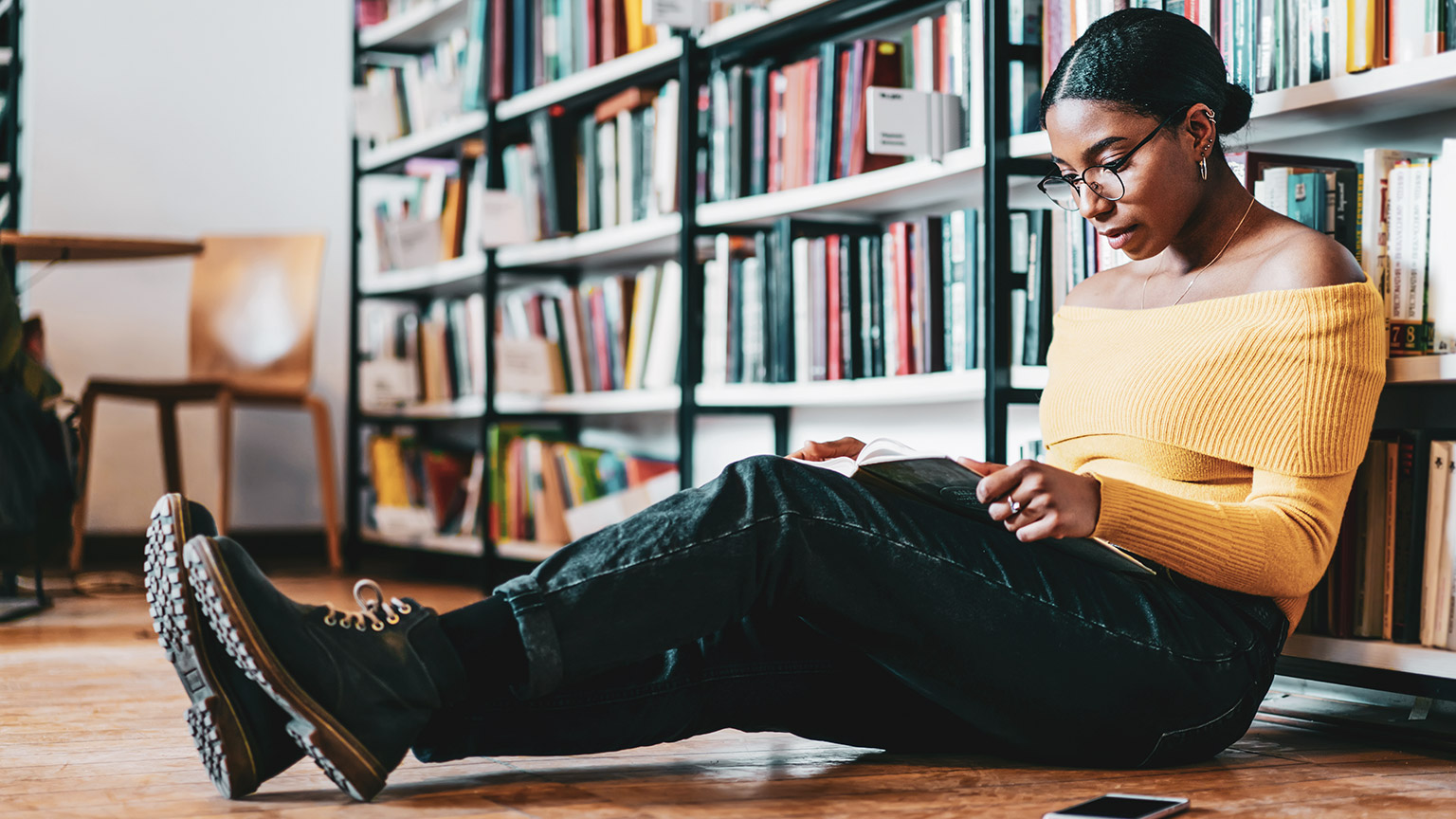 A young designer leaning against a bookshelf in the library, reading a book on Typography