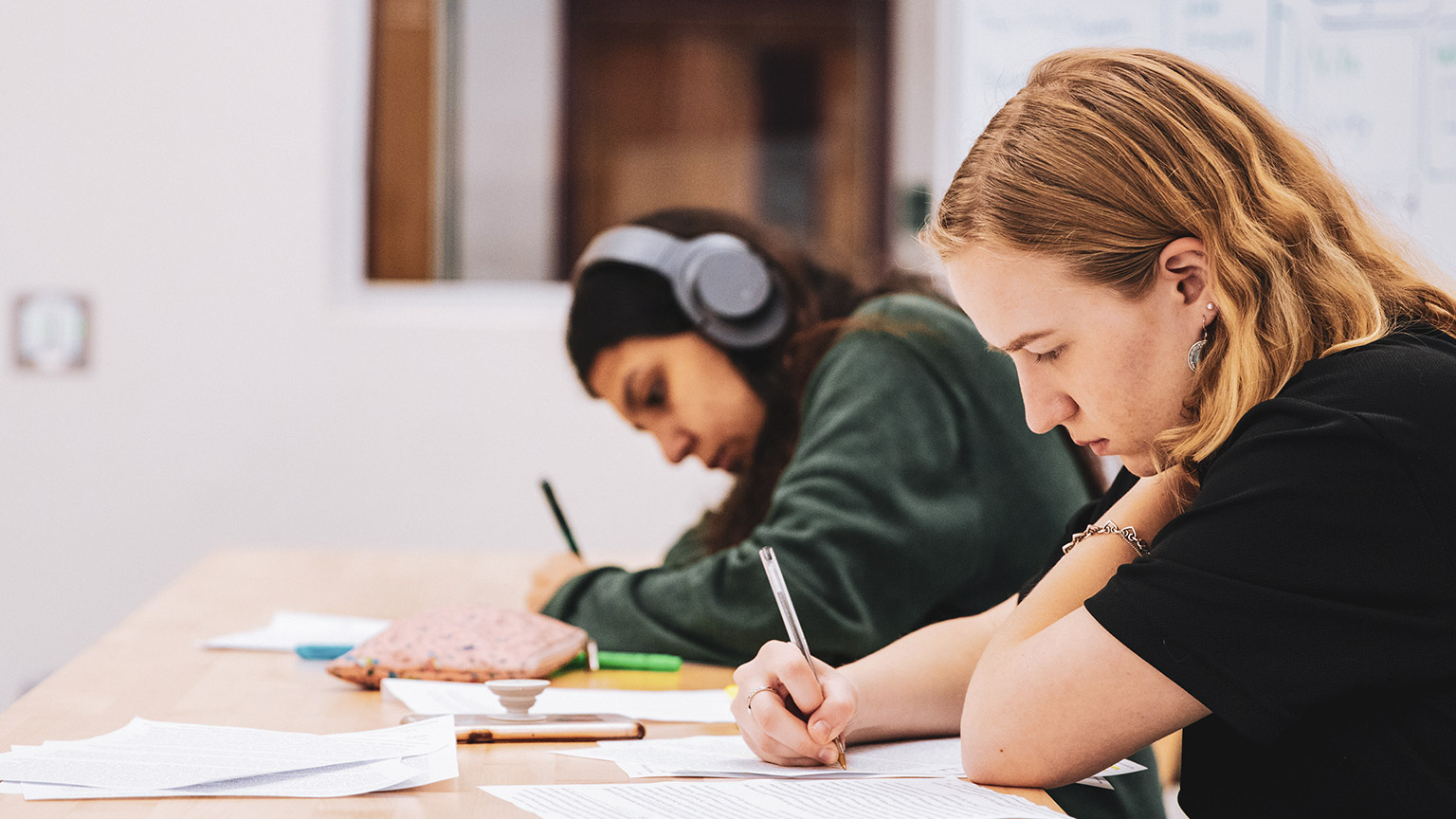 A pair of teenagers seated at their desks, taking down notes in a class