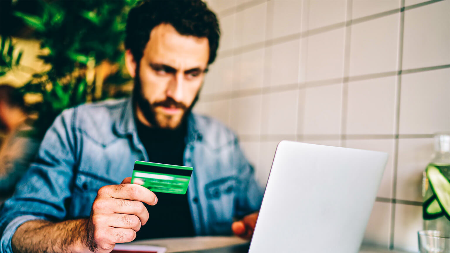 A person seated at a table, holding their credit card while staring at their laptop, considering an online purchase