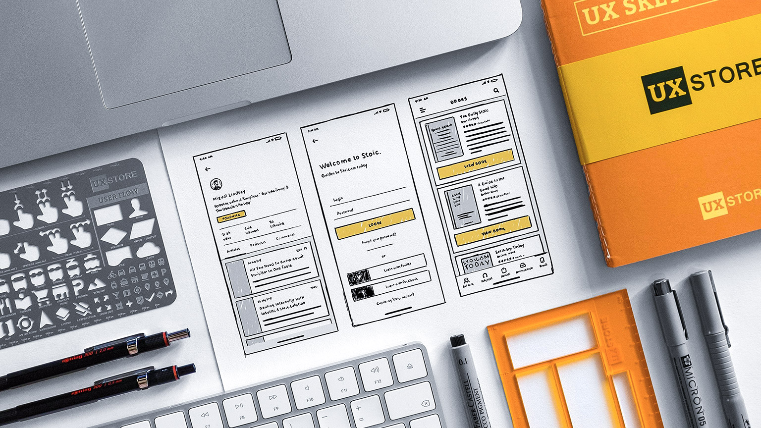 A top down view of a desk with textbook, keyboard, pens and UX thumbnails