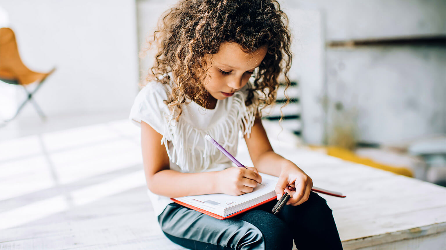 A child seated in their home, learning to write in a small notepad