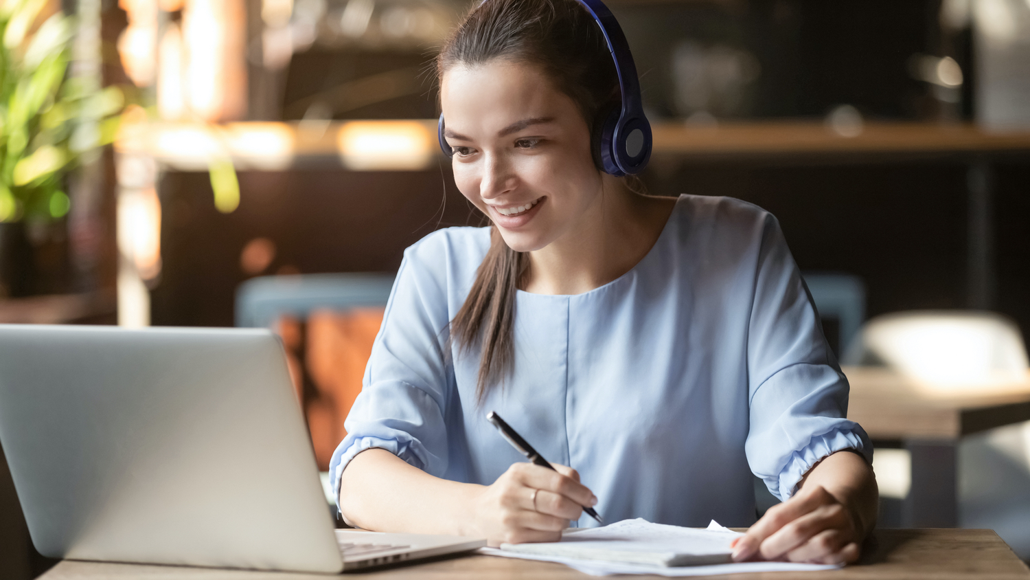 Smiling girl student wear wireless headphone study online with skype teacher, happy young woman learn language listen lecture watch webinar write notes look at laptop sit in cafe, distant education