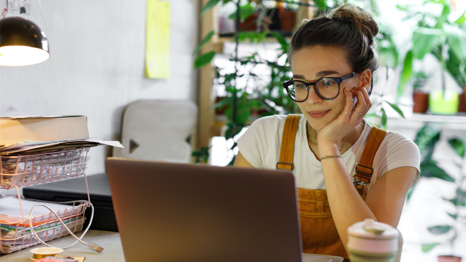 Young female gardener in glasses using laptop, communicates on internet with customer in home garden/greenhouse, reusable coffee/tea mug on table.