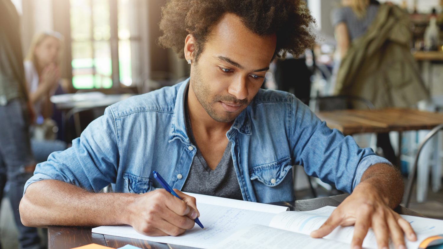 678350452 Portrait of smart hardworking ambitious Afro American student sitting at university canteen, working on research, studying textbook in front of him and writing down important information in copybook