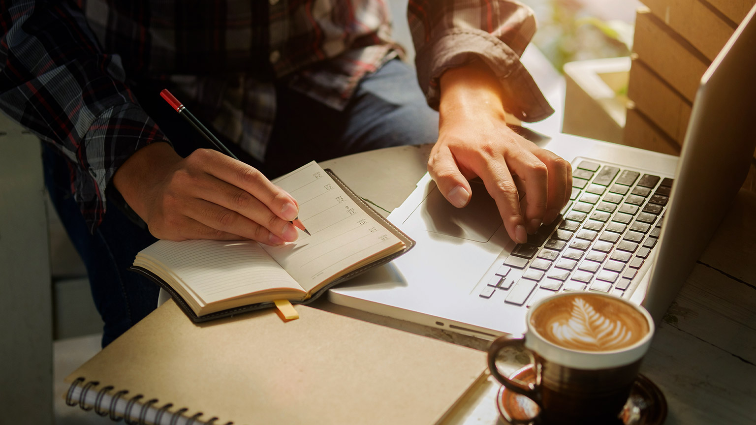 a person with a laptop on one hand, a notebook on the other and a cup of coffee on the table