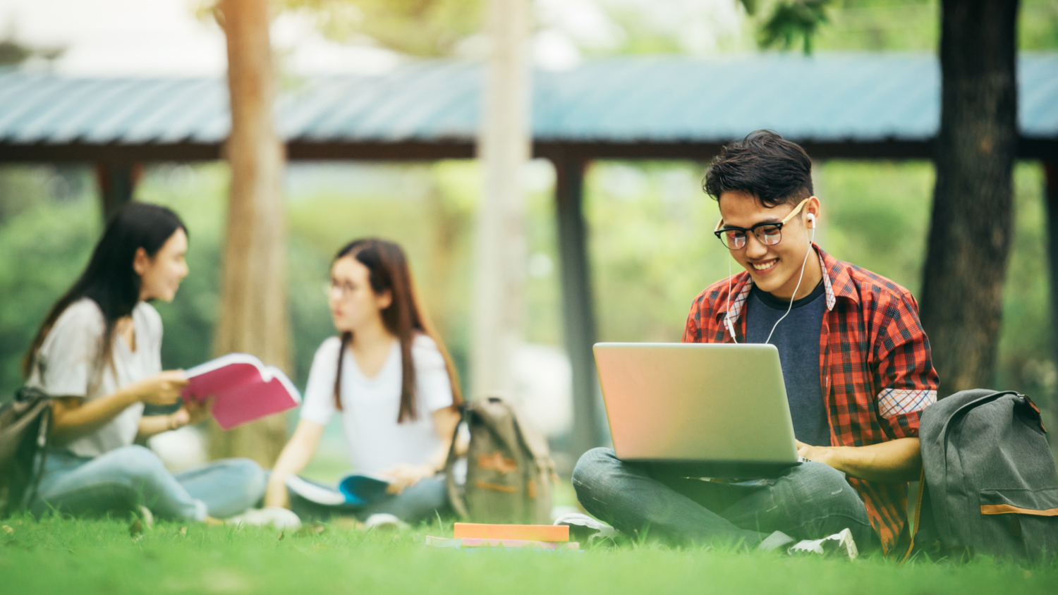 Smiling young male college student sitting on grass studying with a laptop. Two female student in the background having a conversation.