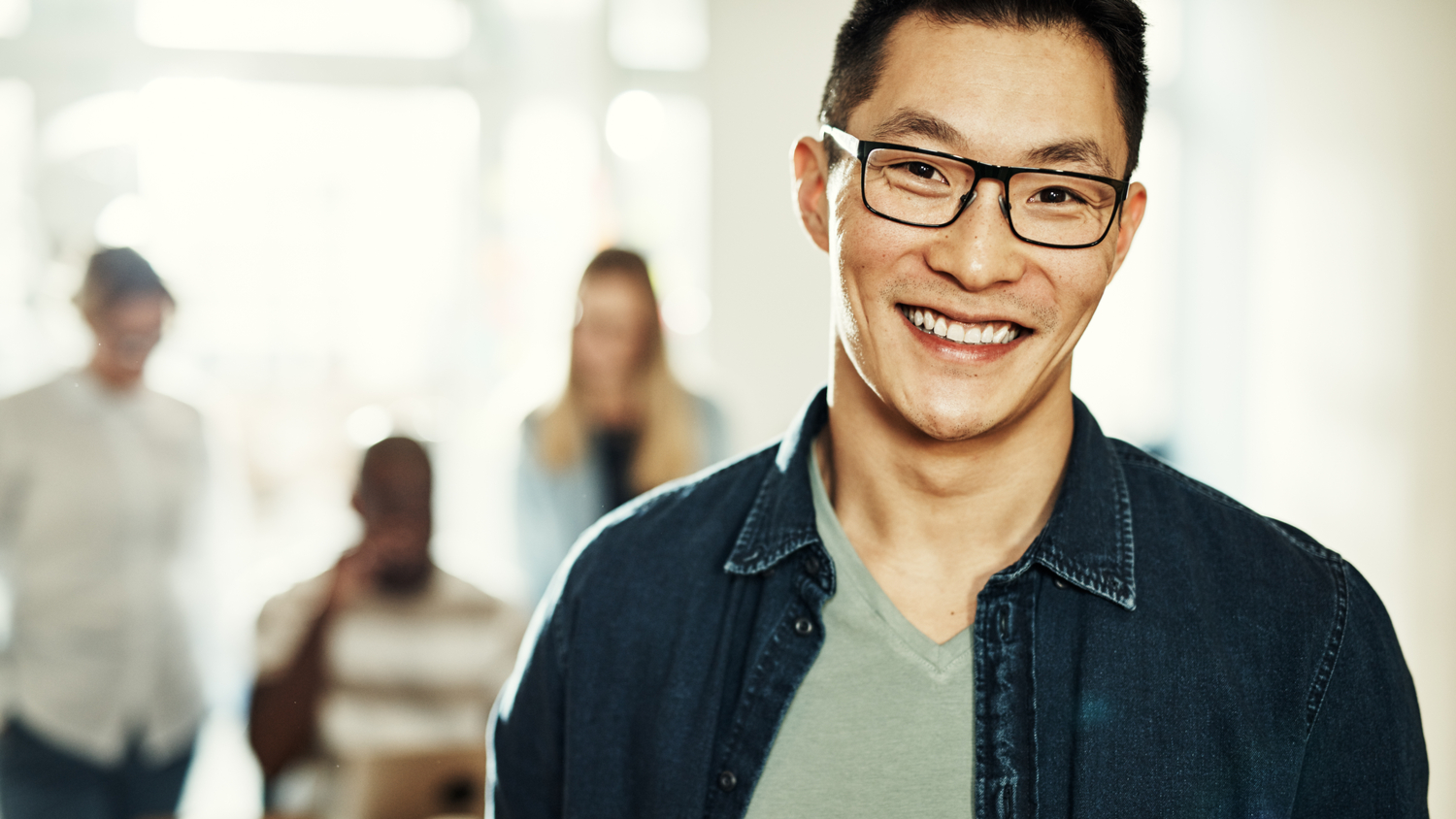 Young Asian manager wearing glasses and smiling confidently while standing in a modern office with colleagues in the background