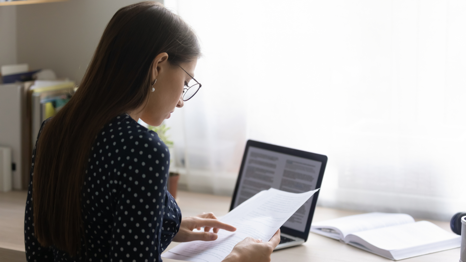 Focused young student in eyeglasses reading paper document and checking policies on her laptop