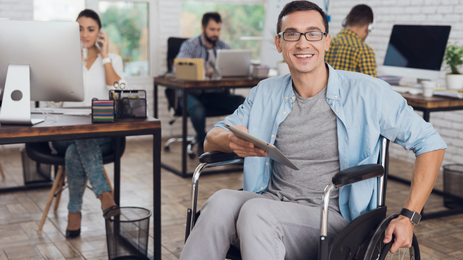 Male employee in the wheelchair working in an office. Smiling at the camera, holding a tablet. His colleagues work nearby at desks