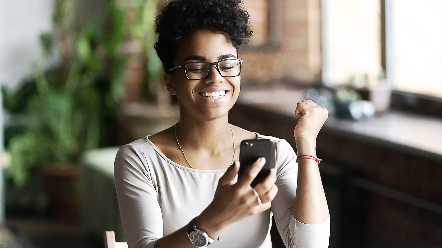 A woman of color raising her fist in joy as she responds to something that she sees on her phone. 