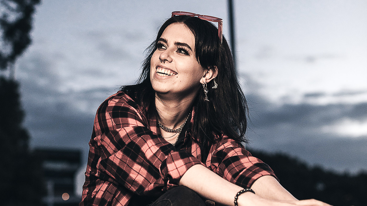 A young woman with glasses on her head, sitting outside at twilight smiling to someone off camera. 