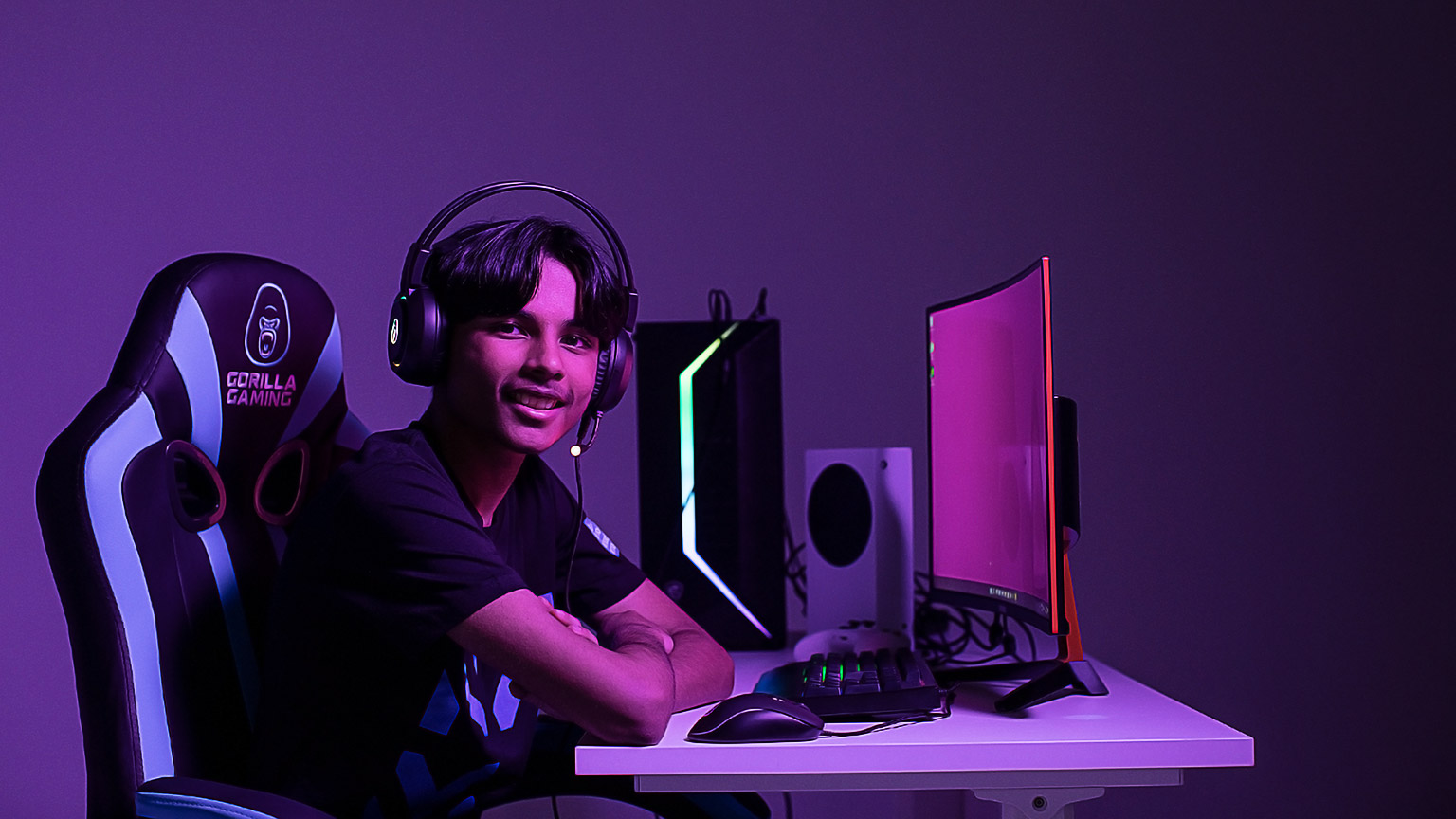 A young man in a room lit in purple, with headphones on, at a desk with many peripherals. He is sitting in a gamer's chair. 