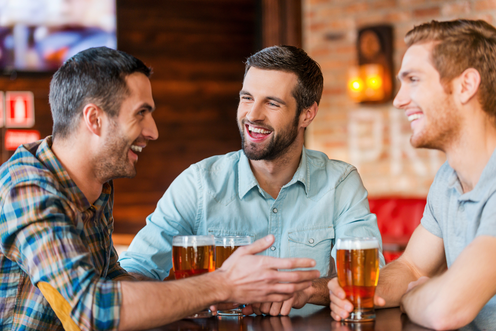 Three happy young Caucasian men in casual wear talking and drinking beer while sitting in bar together