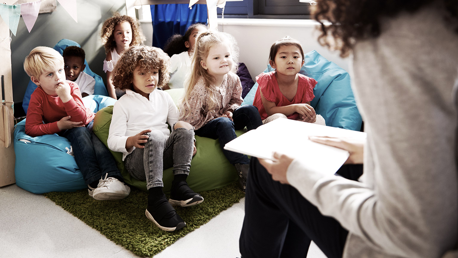 Several children sitting on a matt listening to a teacher reading from a book. 