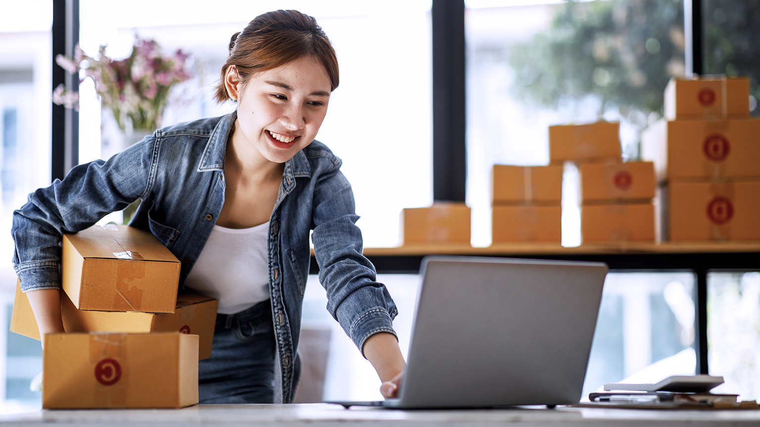 Young woman smiling while holding boxes and working with computer