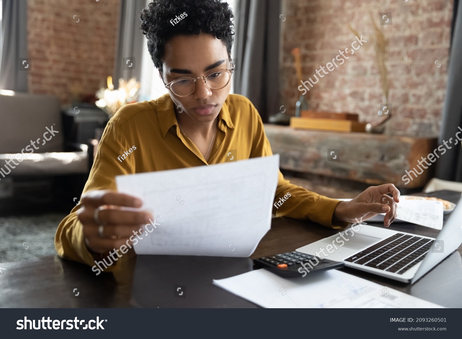 Focused young African American woman in eyeglasses looking through paper documents, managing business affairs, summarizing taxes, planning future investments, accounting alone at home office.