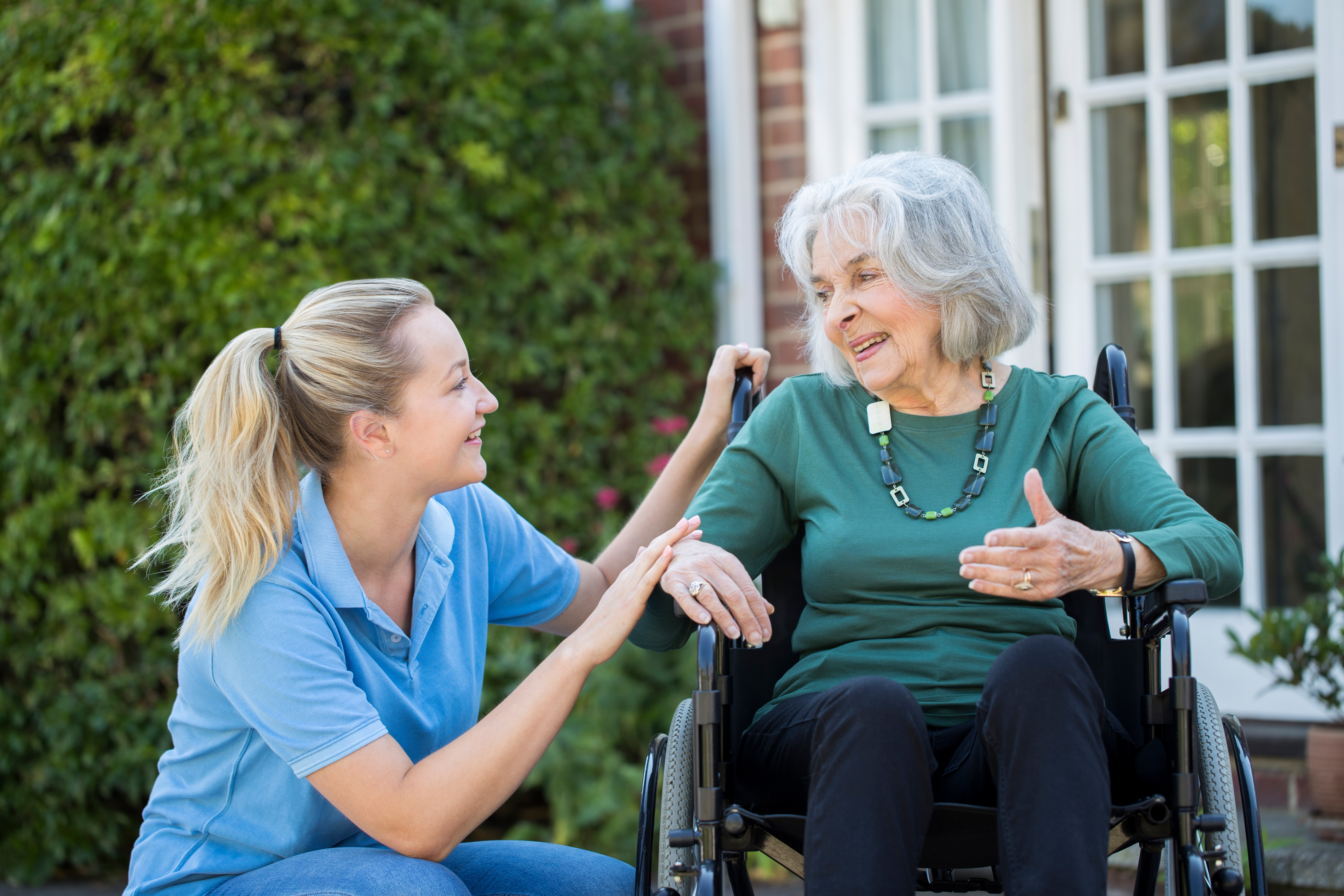 Support worker kneeling beside senior woman in wheelchair outside a home