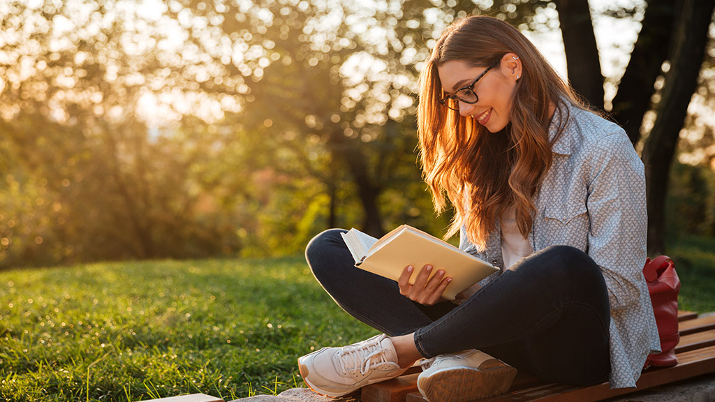 Side view of pleased brunette woman in eyeglasses sitting on bench and reading book in park