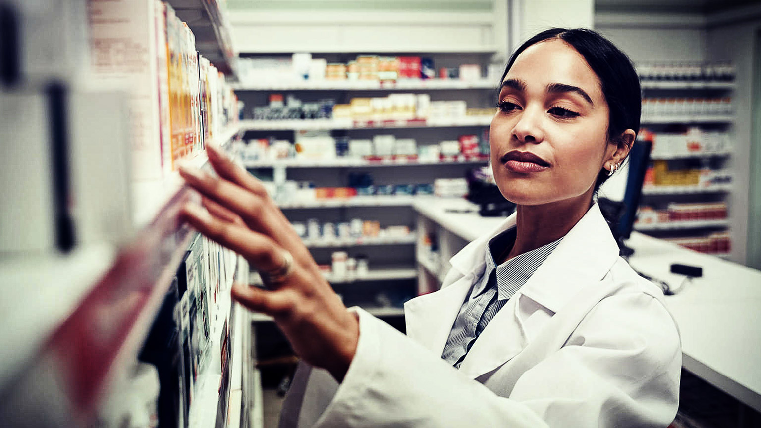 Young woman working in pharmacy looking for medicine in shelf standing behind counter