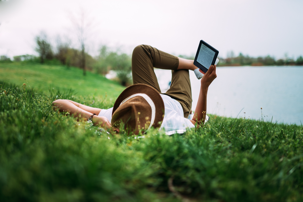 Person laying on the grass outdoors. Reading on a tablet next to lake.