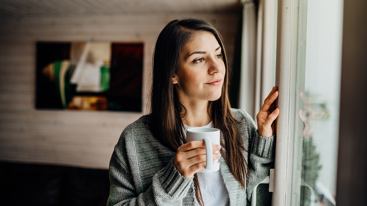 A person gazing out a window while holding a cup of coffee and contemplating matters of mental health