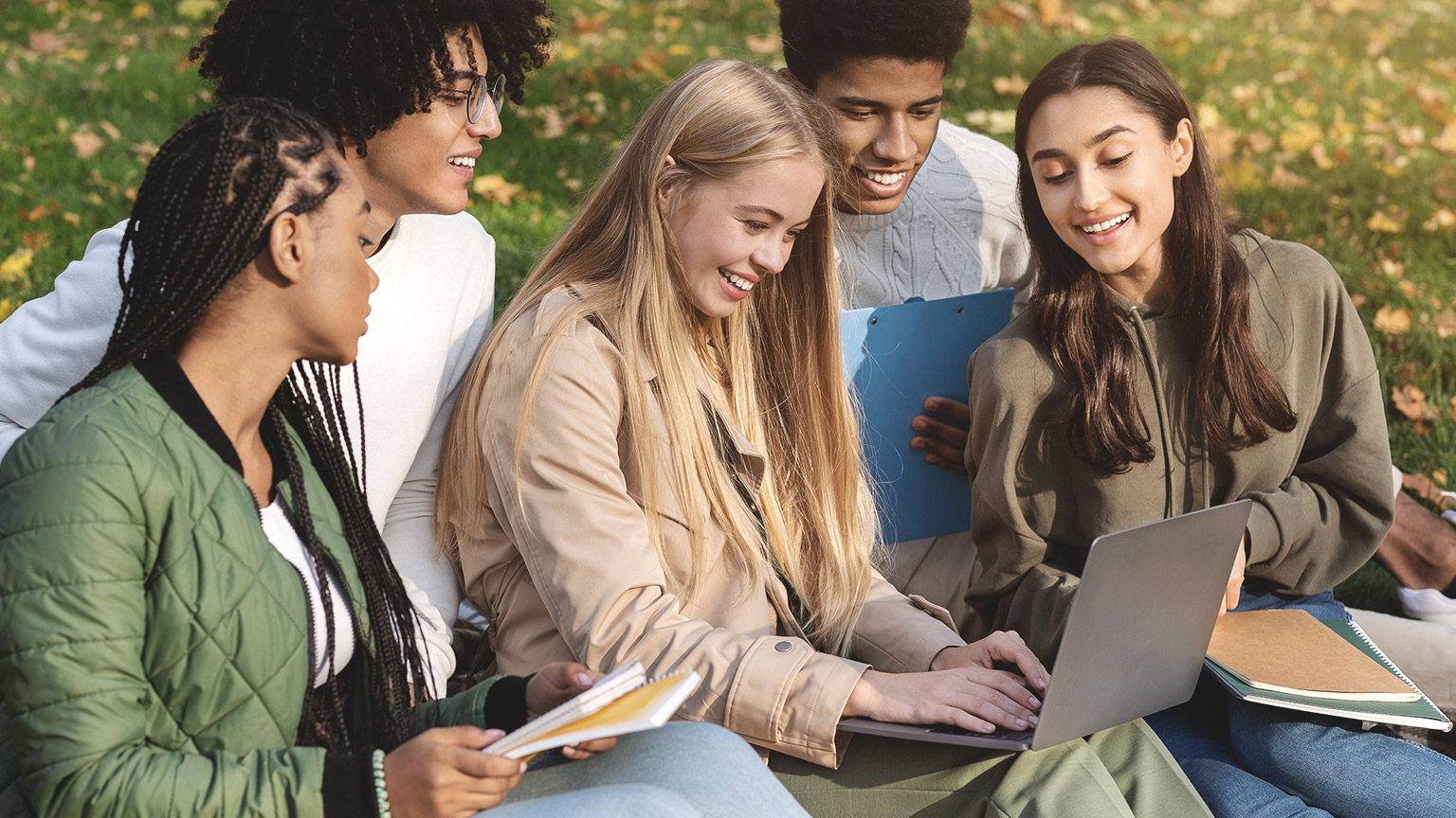 Group of students looking at a computer while sitting on a grass outdoor