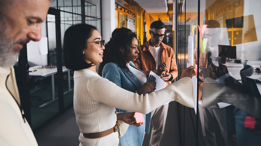 Side view of group of multiethnic coworkers in casual clothes standing near glass wall and attaching sticky notes