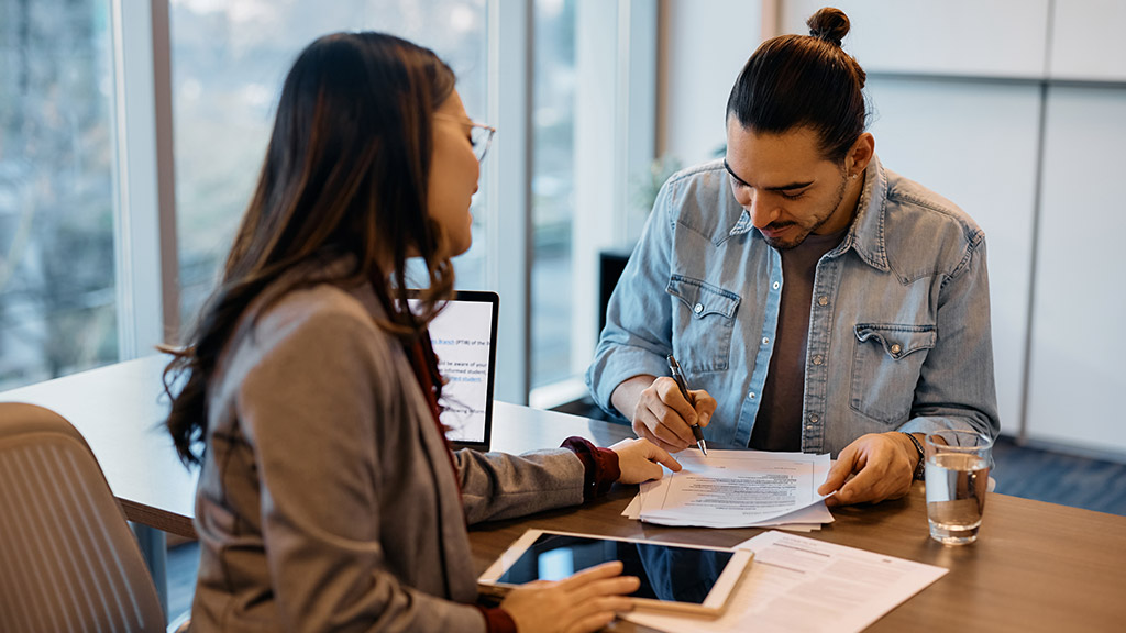 Happy Hispanic man signing paperwork while having meeting with insurance agent in the office