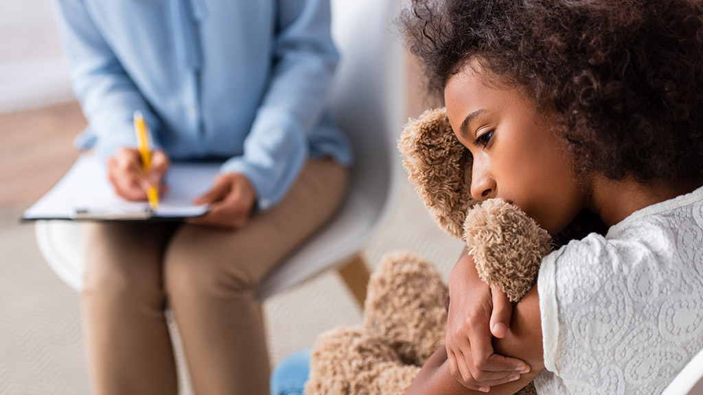 Upset african american girl with autism hugging teddy bear during consultation with blurred psychologist on background