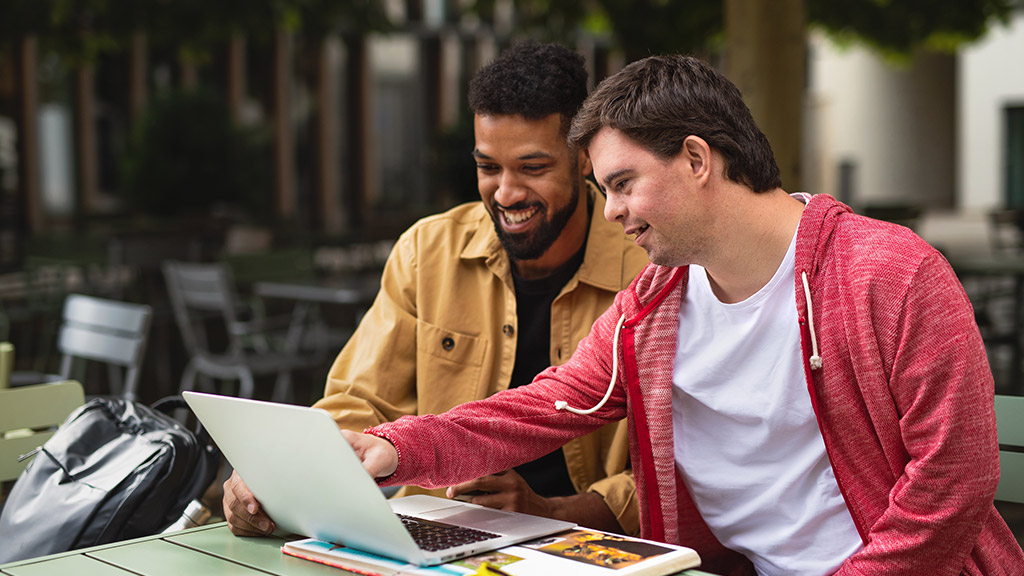 Young man with Down syndrome with his mentoring friend sitting outdoors in cafe using laptop.