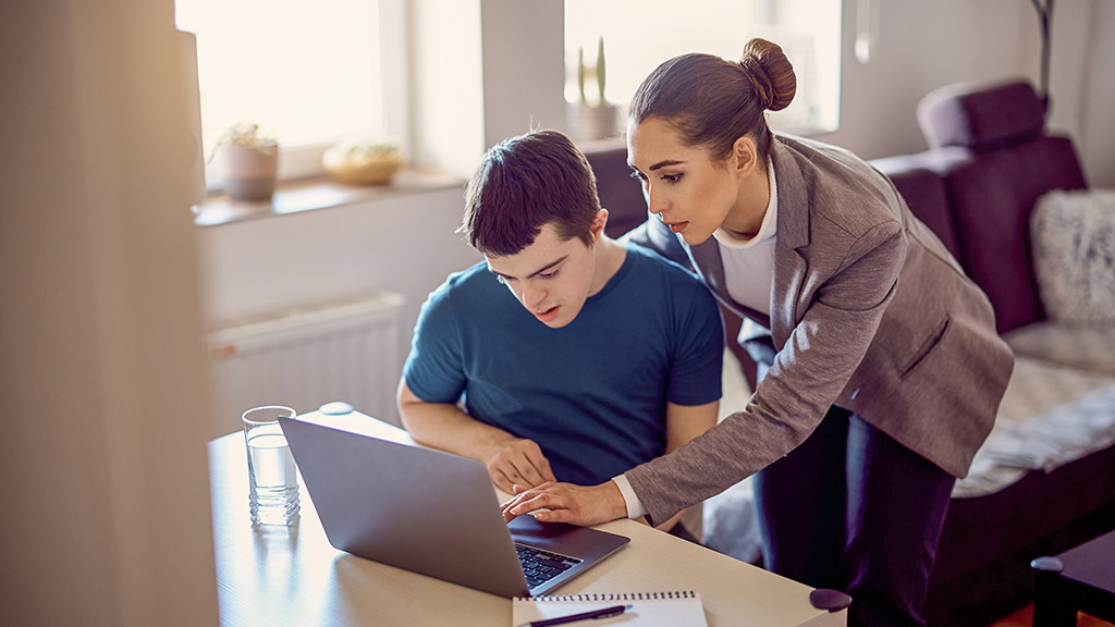 young man with down syndrome learning to use laptop with help of his tutor at home