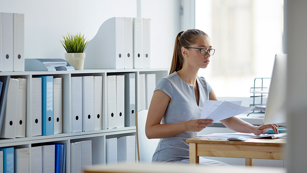 female office worker at computer desk sorting documentation and data alone in light spacious room