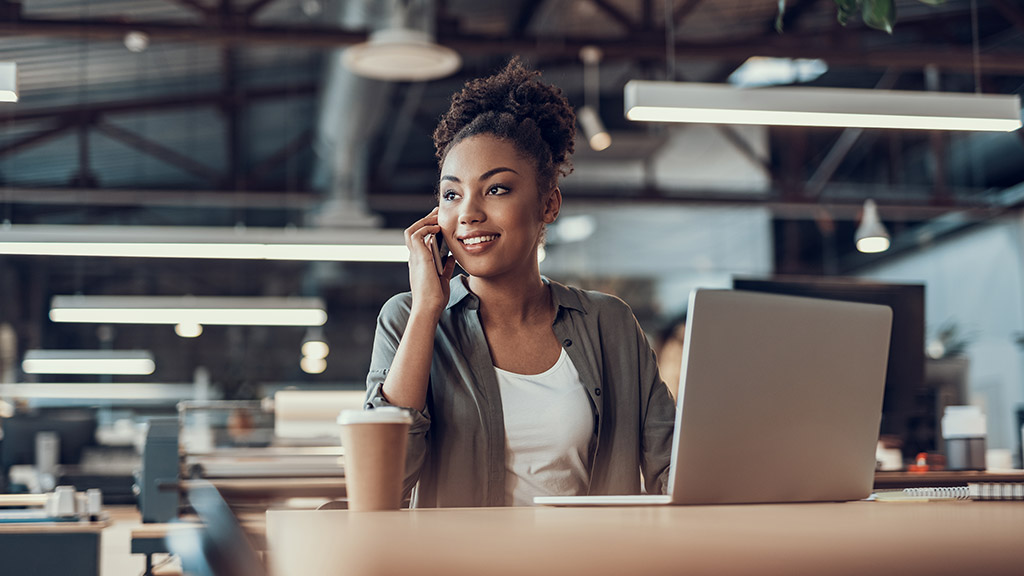 charming young lady having phone conversation while sitting at office table with notebook and cup of coffee