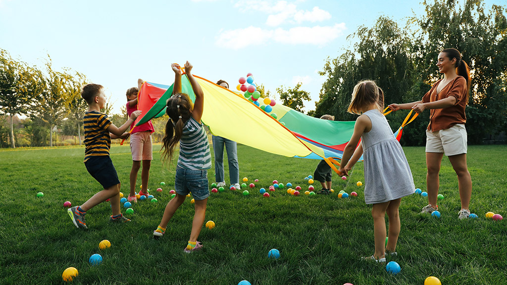 Group of children and teacher playing with rainbow playground parachute on green grass