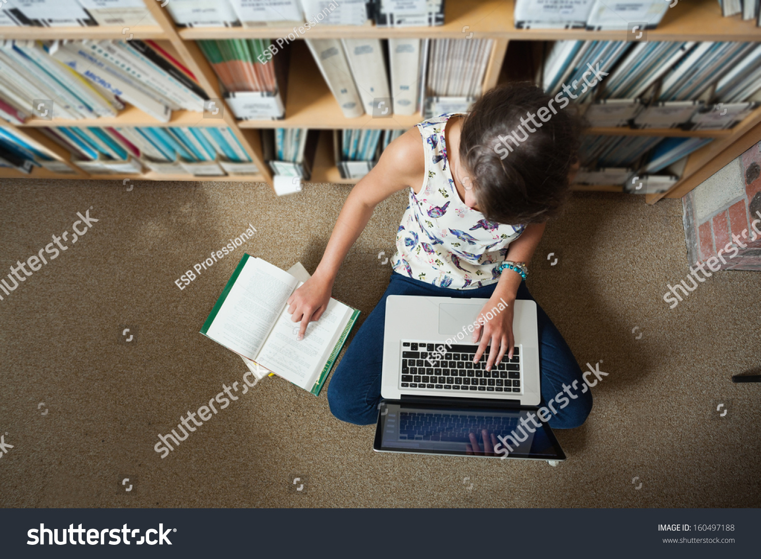 High angle view of a female student sitting against bookshelf with laptop on the library floor