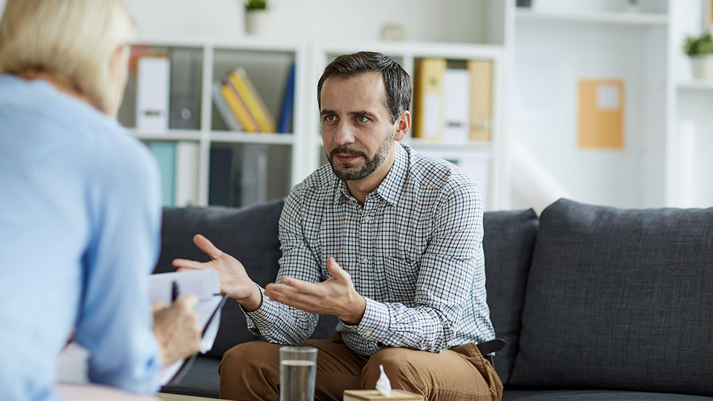 Tense middle aged man in casualwear explaining his trouble to psychologist while sitting on couch in front of her