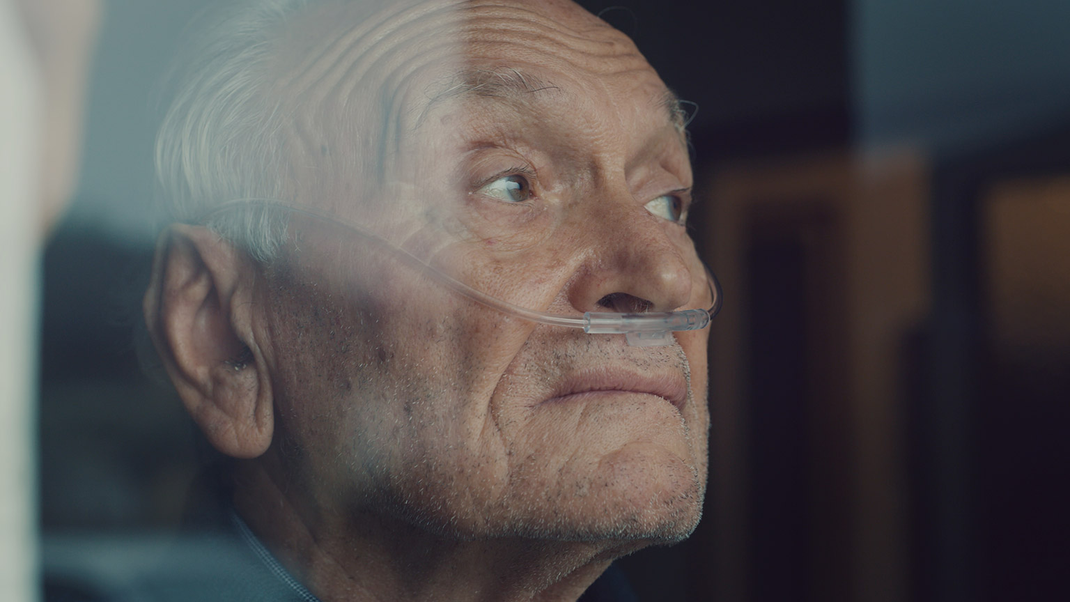 An older person with breathing tube looking out a window