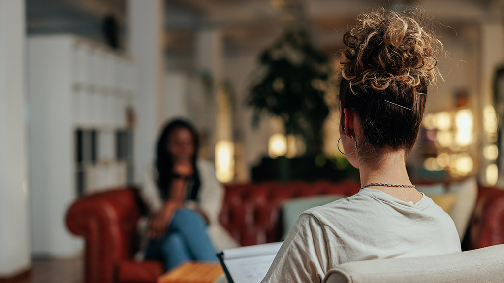 Focus on female psychiatrist writing information of african female patient while sitting on sofa and talking about his illness