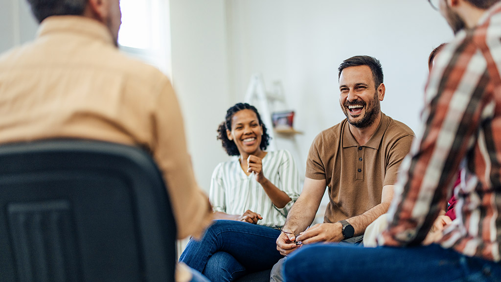 Focus on the smiling man, talking with people of all ages, during the group therapy