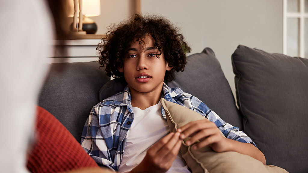 Cute teenage boy sitting on a sofa with pillows and gesturing while talking.