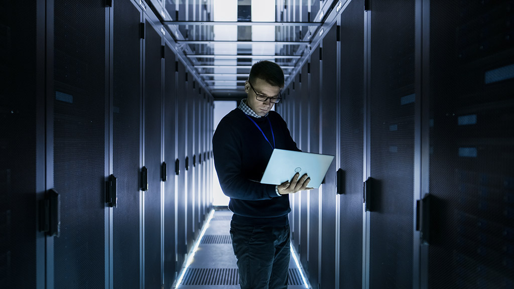 male it engineer works on a laptop in front of server cabinet at a big data center